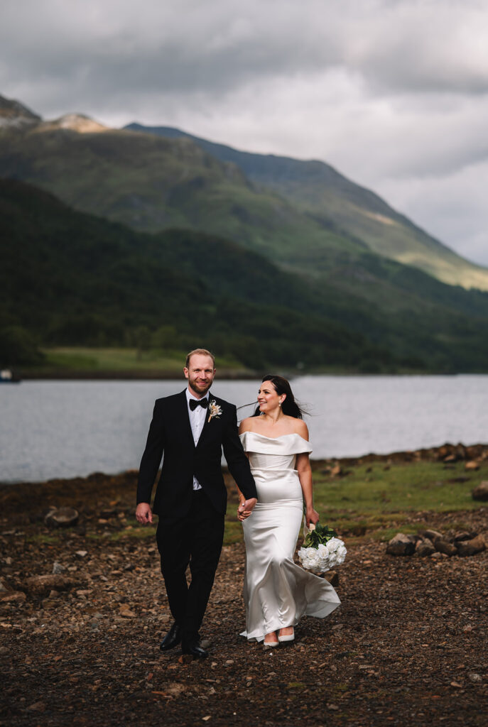 Bride and groom walking by lakeside with mountains in the background.