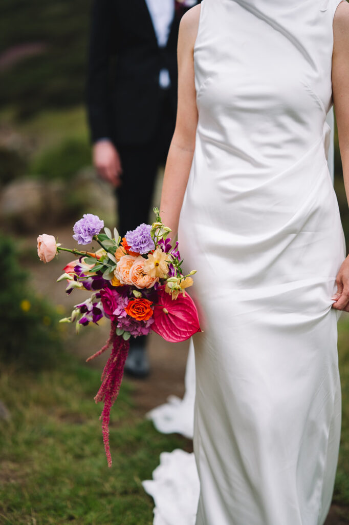 Bride holding vibrant bouquet of colorful flowers during outdoor wedding with groom blurred in background.