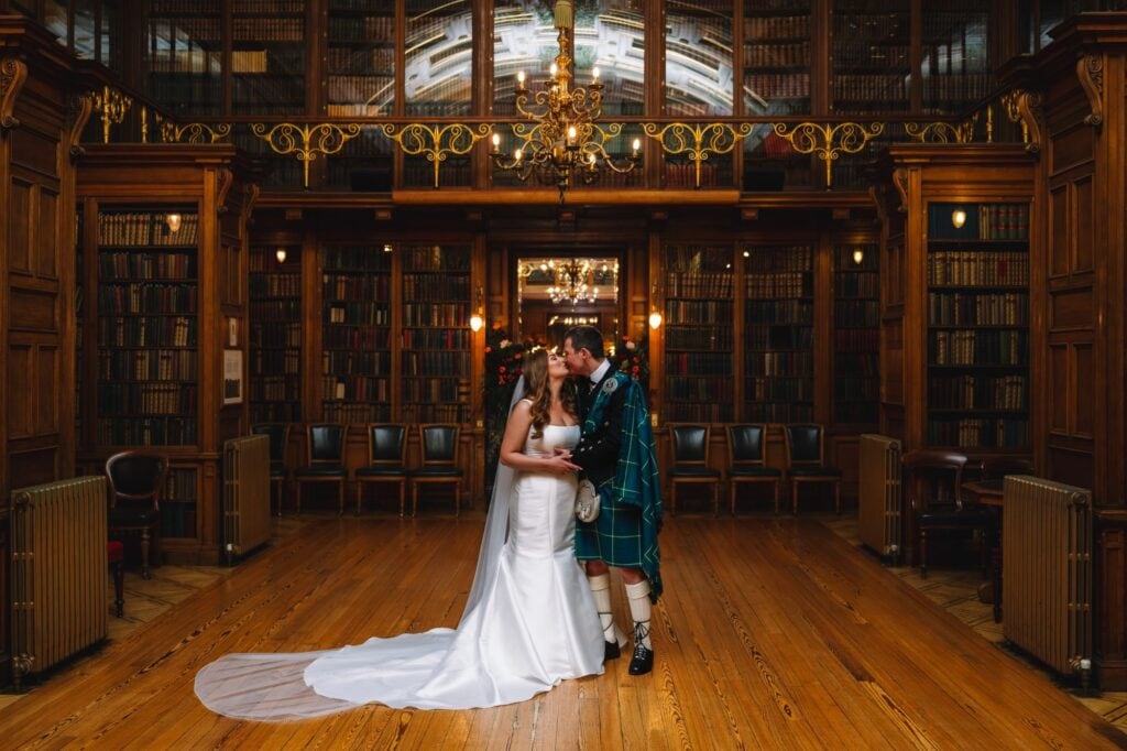 A wedding ceremony in a grand library at the Royal College of Physicians in Edinburgh features tall bookshelves. The bride in a white gown and the groom in a kilt stand before an officiant. Guests sit on wooden chairs, while a chandelier and large mirror elegantly capture this scene in Scotland.