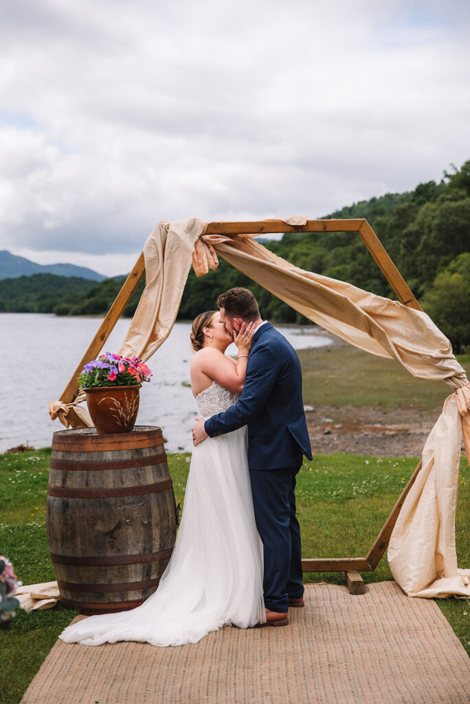 Couple kissing under wooden arch by lakeside ceremony setup.