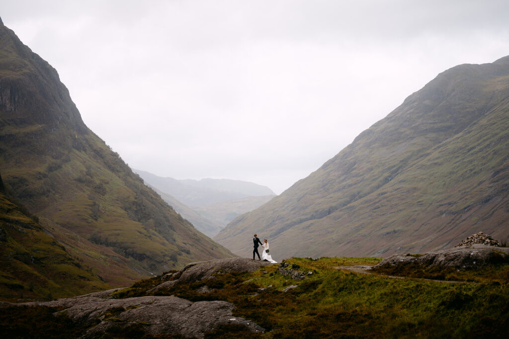 Couple standing on rocky hilltop in misty mountain landscape.