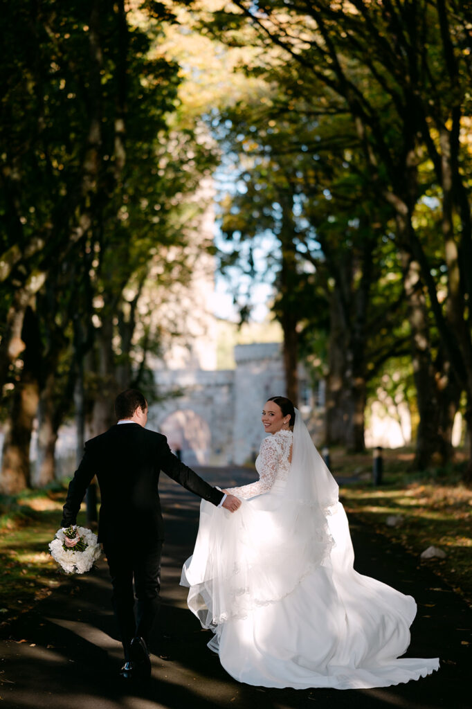 Bride and groom walking hand in hand through tree-lined path.