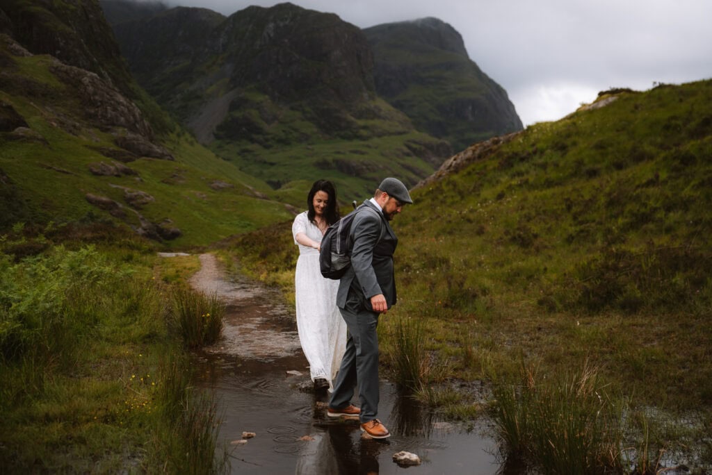 A man and woman walk through a puddle on a mountain path in Glencoe. The man, wearing a suit and flat cap, leads while the woman in a white dress follows. Lush green hills and cloudy skies envelop them, setting an idyllic scene for their elopement in the heart of Scotland.