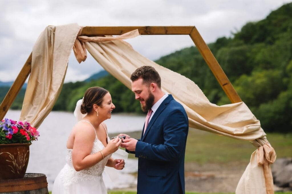 Couple exchanging rings under wooden arch by water