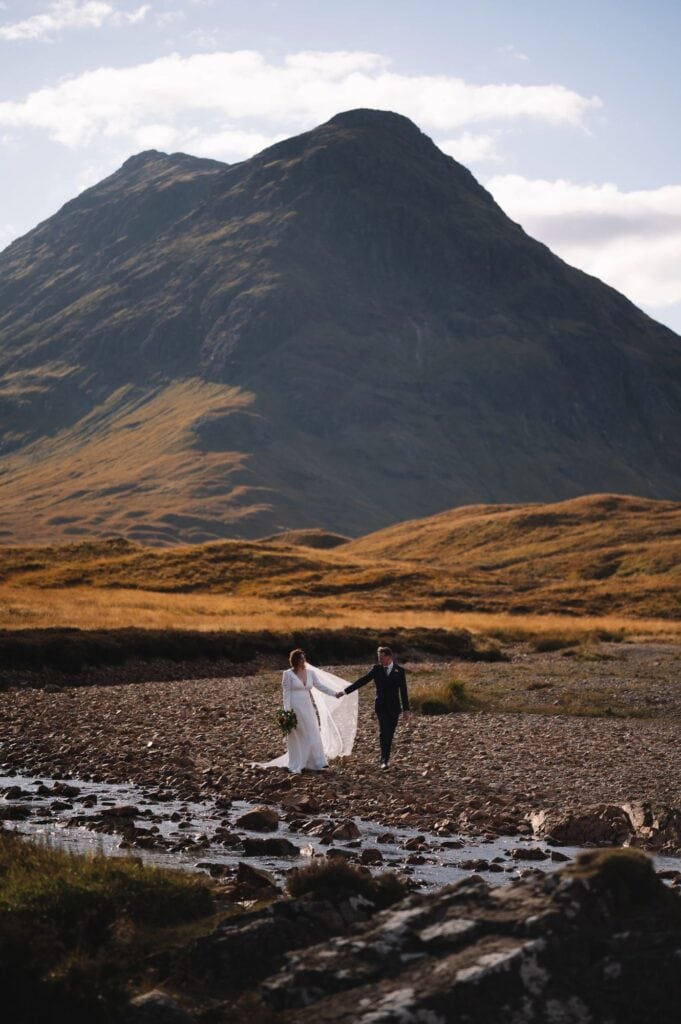 A couple in wedding attire walks hand in hand across a rocky stream in an expansive, mountainous landscape in Glencoe, Scotland. The bride holds a bouquet and the groom is in a dark suit. A tall, grassy mountain rises in the background under a partly cloudy sky.