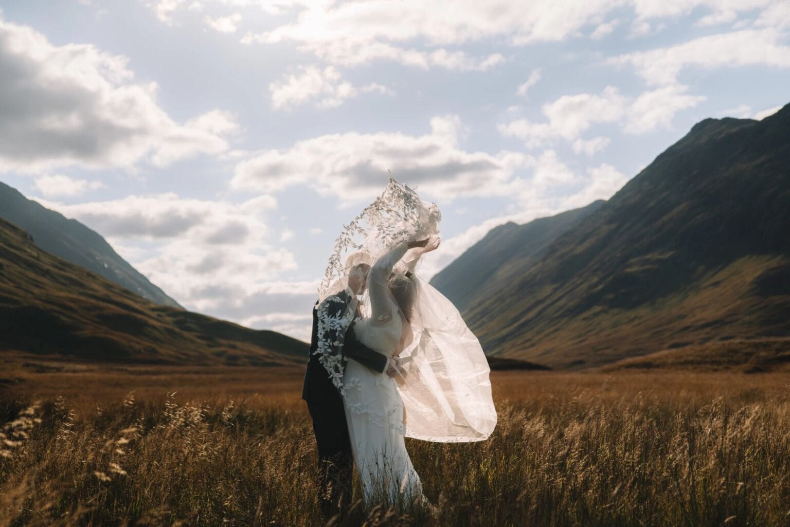 A couple in wedding attire joyfully embraces in a grassy field at Glencoe, Scotland, surrounded by scenic mountains under a partly cloudy sky. The brides veil flows in the wind, capturing a sense of freedom and elopement celebration.