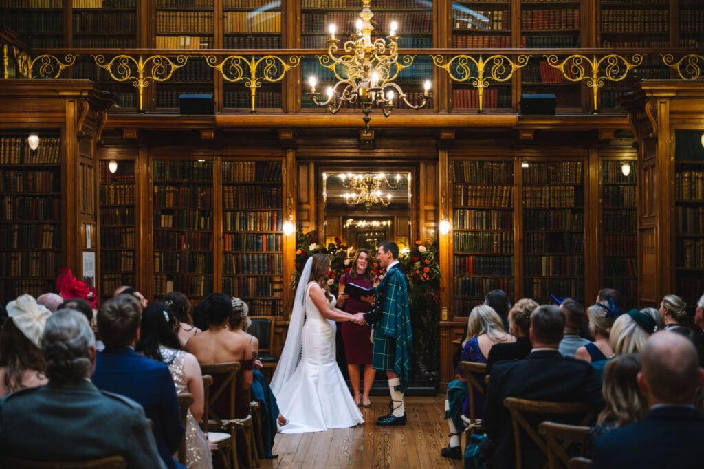 A couple stands at the altar in a library-themed venue, exchanging vows at their Edinburgh wedding. The bride wears a long white dress with a veil, and the groom is in a kilt. Guests sit in rows surrounded by bookshelves, while a chandelier hangs overhead, adding elegance to this Scottish celebration.
