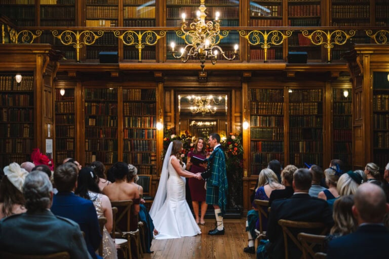 A couple stands at the altar in a library-themed venue, exchanging vows at their Edinburgh wedding. The bride wears a long white dress with a veil, and the groom is in a kilt. Guests sit in rows surrounded by bookshelves, while a chandelier hangs overhead, adding elegance to this Scottish celebration.