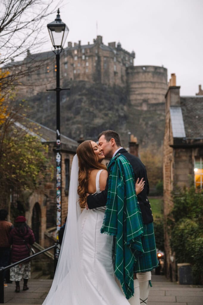 A bride and groom embrace on the Vennels cobblestone street, with the groom wearing a green tartan over his kilt. The bride has a long veil and white dress. In the background, Edinburgh Castle perches atop a rocky hill. The scene is overcast and autumnal.