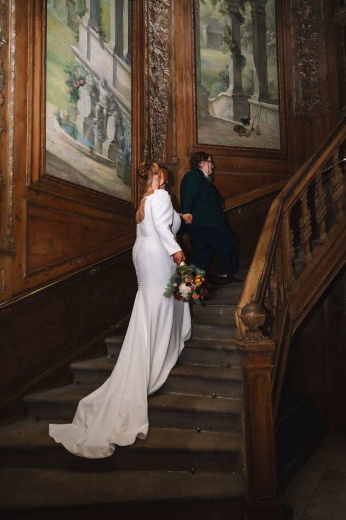 Bride ascending staircase with groom in elegant historic interior.