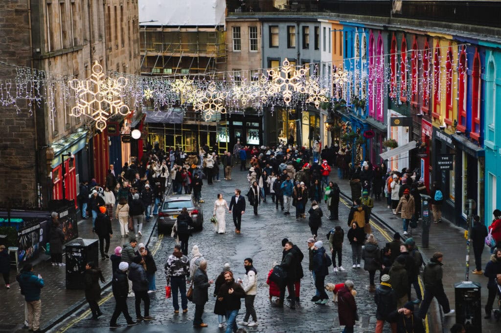 A bustling Edinburgh city street (Victoria Street) scene with people walking on wet cobblestones. Colorful buildings line the vibrant Victoria Street, adorned with hanging festive lights shaped like snowflakes. The atmosphere is lively and captivating, echoing the charm of Holyrood Park nearby.