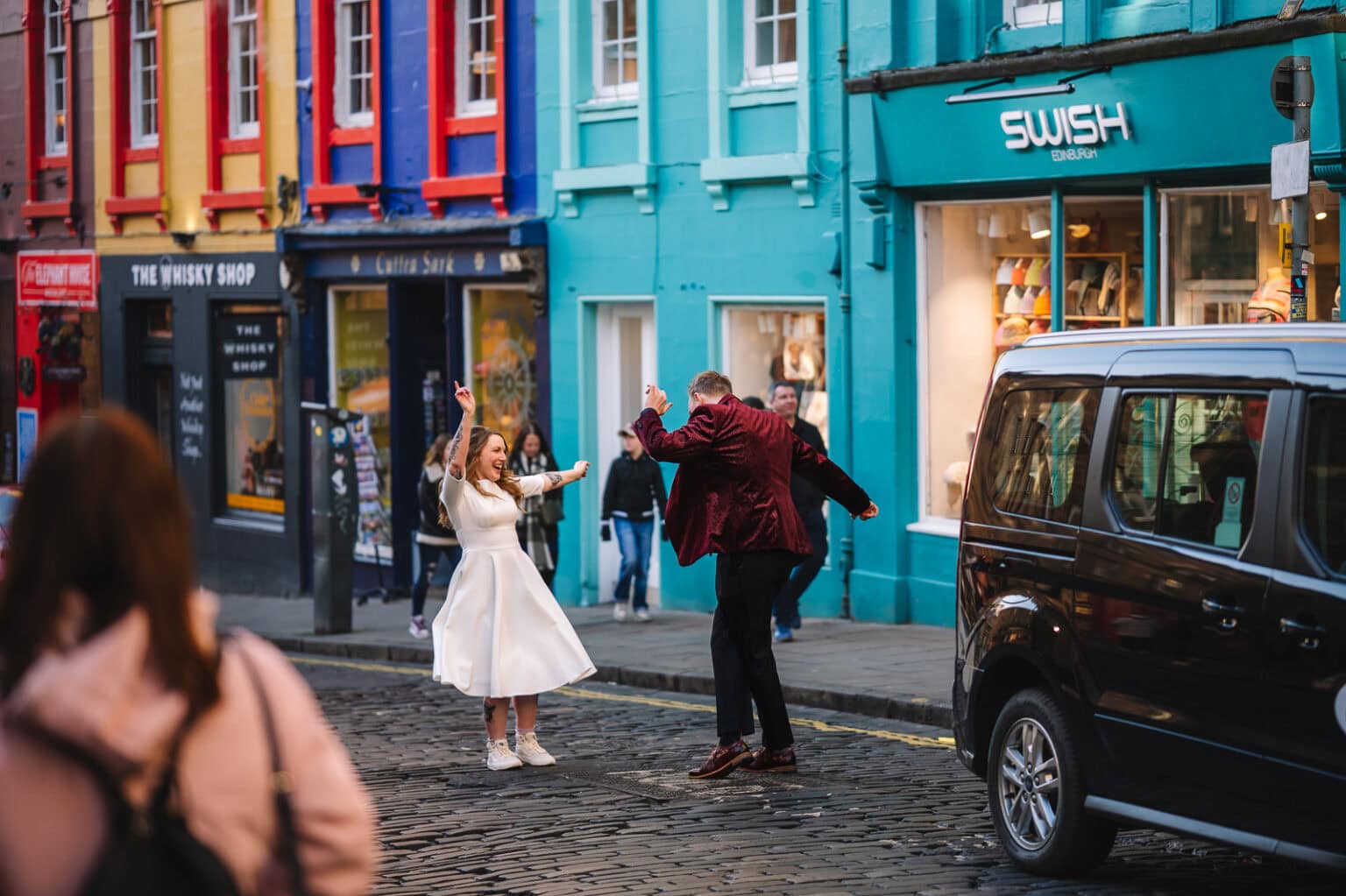 A couple joyfully dances on a cobblestone street in Edinburgh, lined with colorful buildings. The woman is in a white dress, and the man wears a red jacket. Near a quaint tattoo shop, a black car is parked with a few people celebrating their micro wedding in the background.