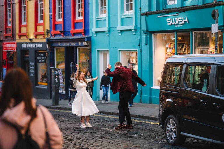 A couple joyfully dances on a cobblestone street in Edinburgh, lined with colorful buildings. The woman is in a white dress, and the man wears a red jacket. Near a quaint tattoo shop, a black car is parked with a few people celebrating their micro wedding in the background.