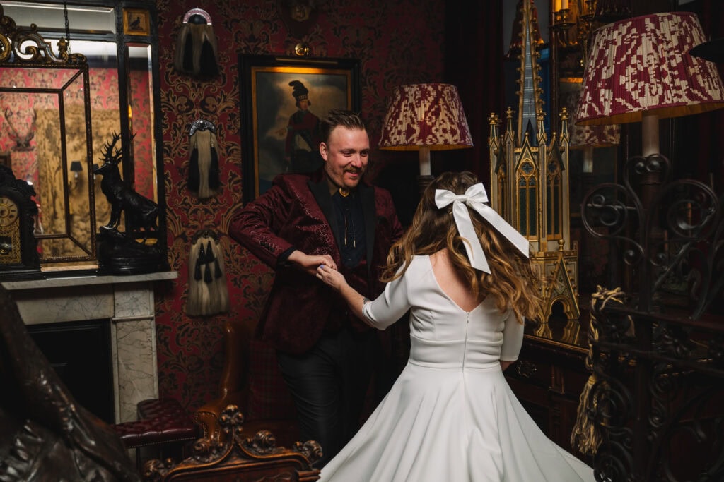 A couple dances joyfully in a richly decorated room reminiscent of an Edinburgh micro wedding. The man wears a dark red velvet jacket, while the woman dazzles in a white dress with a bow in her hair. Ornate wallpaper and eclectic decor surround them, with a painting and decorative items visible.