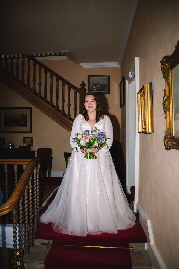Bride standing on staircase holding bouquet in elegant interior.