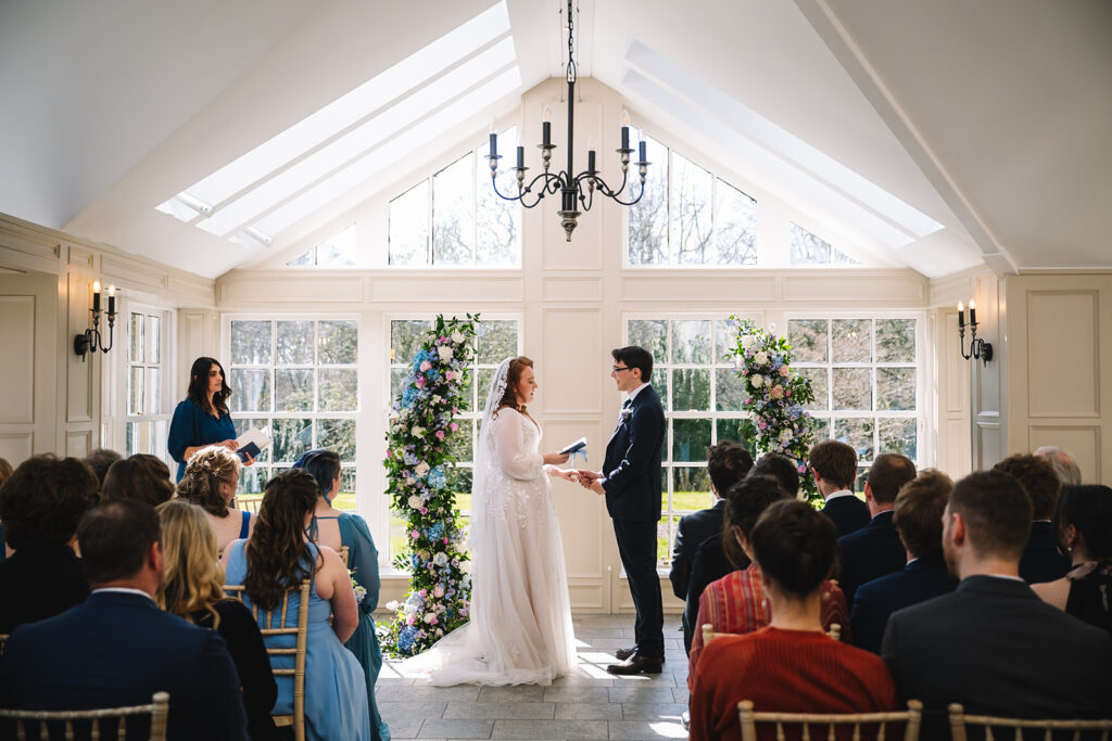 Indoor ceremony with couple exchanging vows in bright glass-roofed venue.