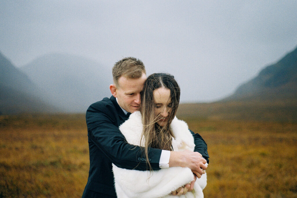 Groom hugging bride from behind during emotional outdoor moment.