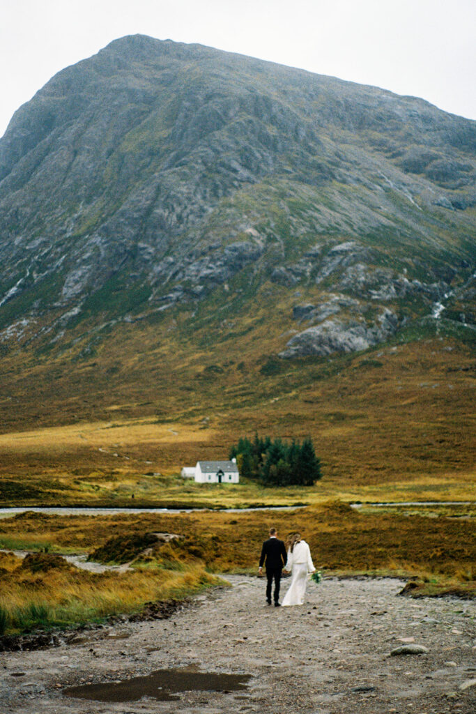 Couple walking through open Highland landscape with small white cottage nearby.