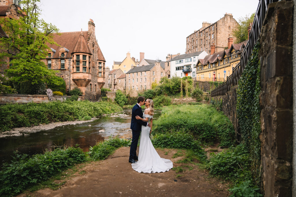 Couple standing by river with historic buildings behind.