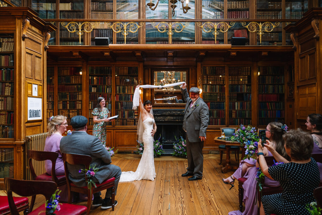 bride and groom standing during ceremony inside library with guests.