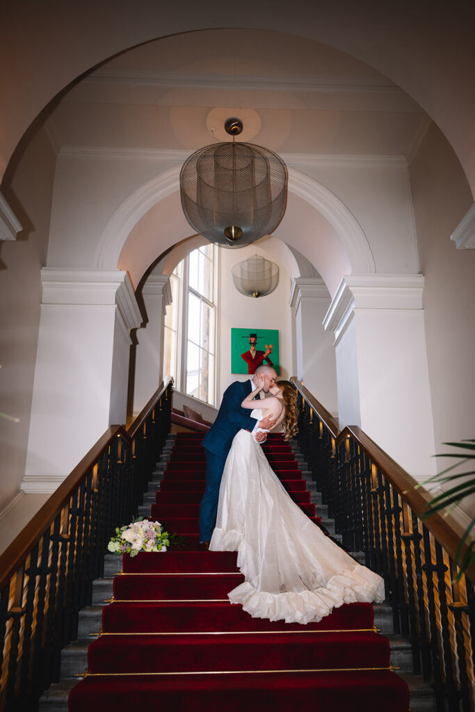 Bride standing on grand staircase with groom in elegant interior.