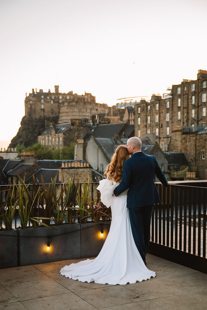 Couple embracing on rooftop with Edinburgh Castle in background.