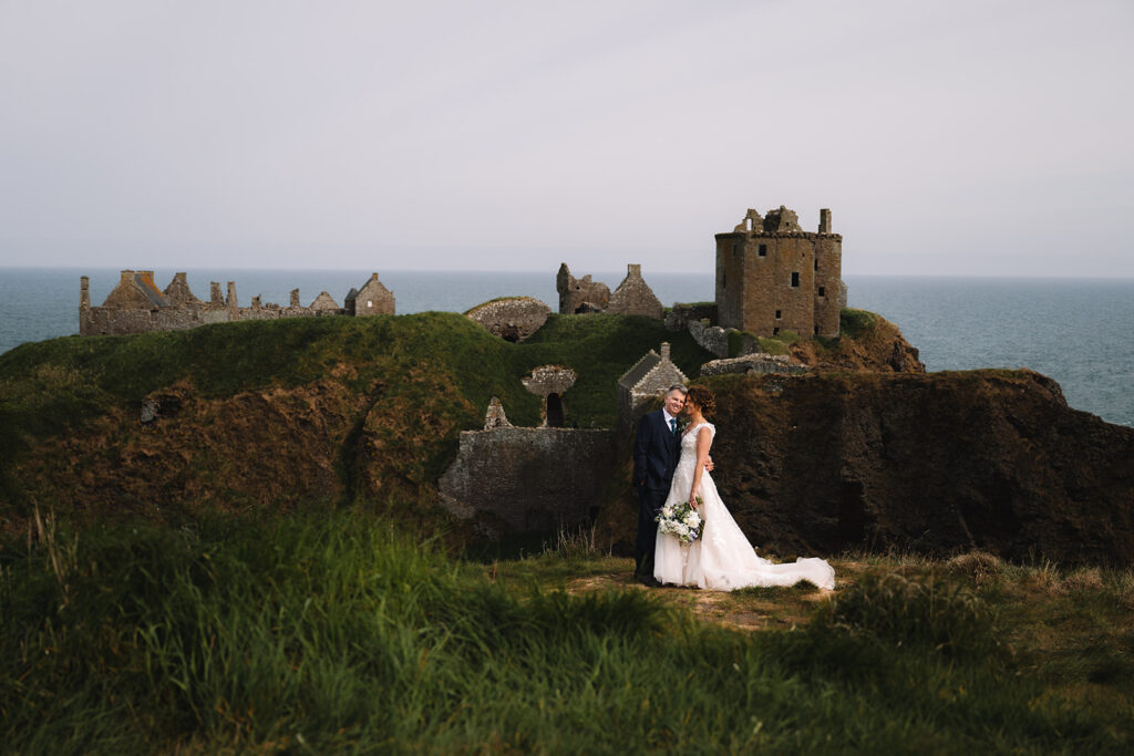 Bride and groom embracing near castle ruins with ocean backdrop.