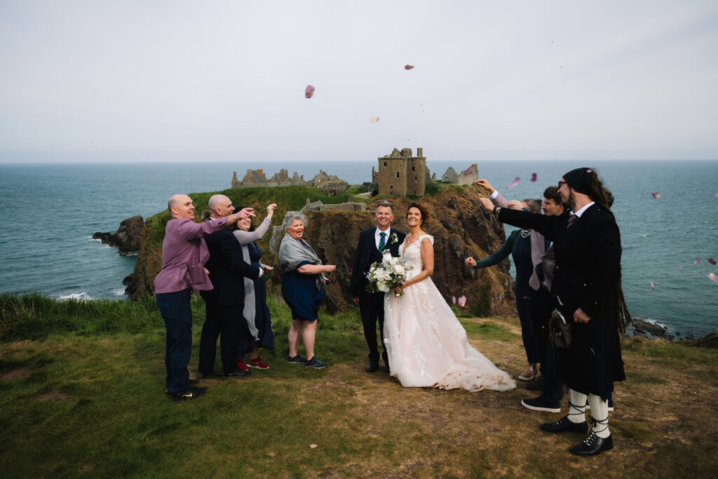 Wedding party celebrating with confetti on cliff edge near castle.