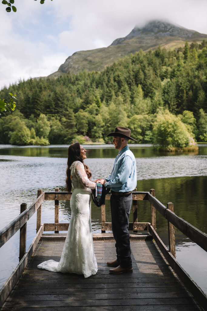 Couple holding hands on dock surrounded by water and trees.