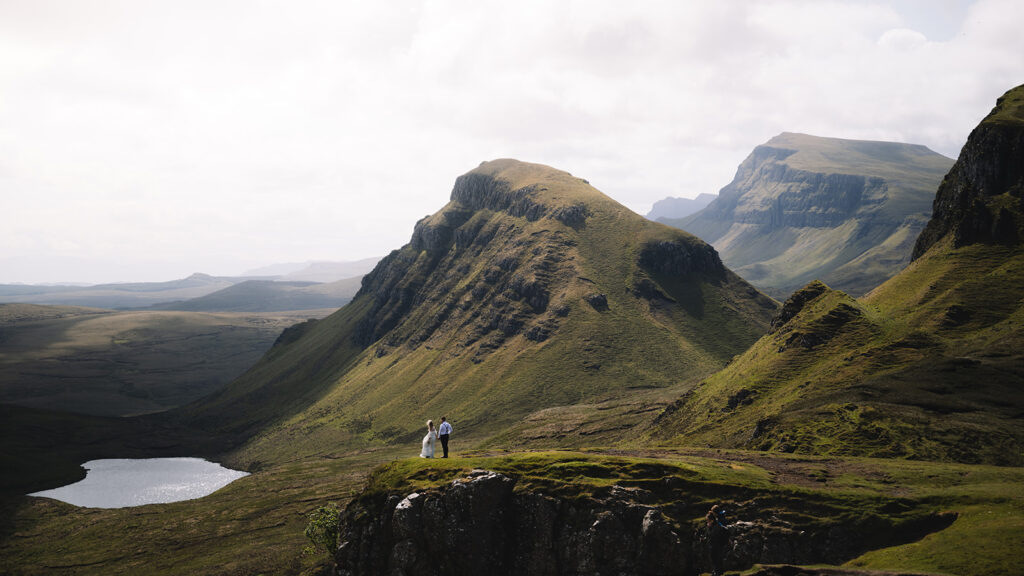 Couple standing together on ridge with layered mountains behind.