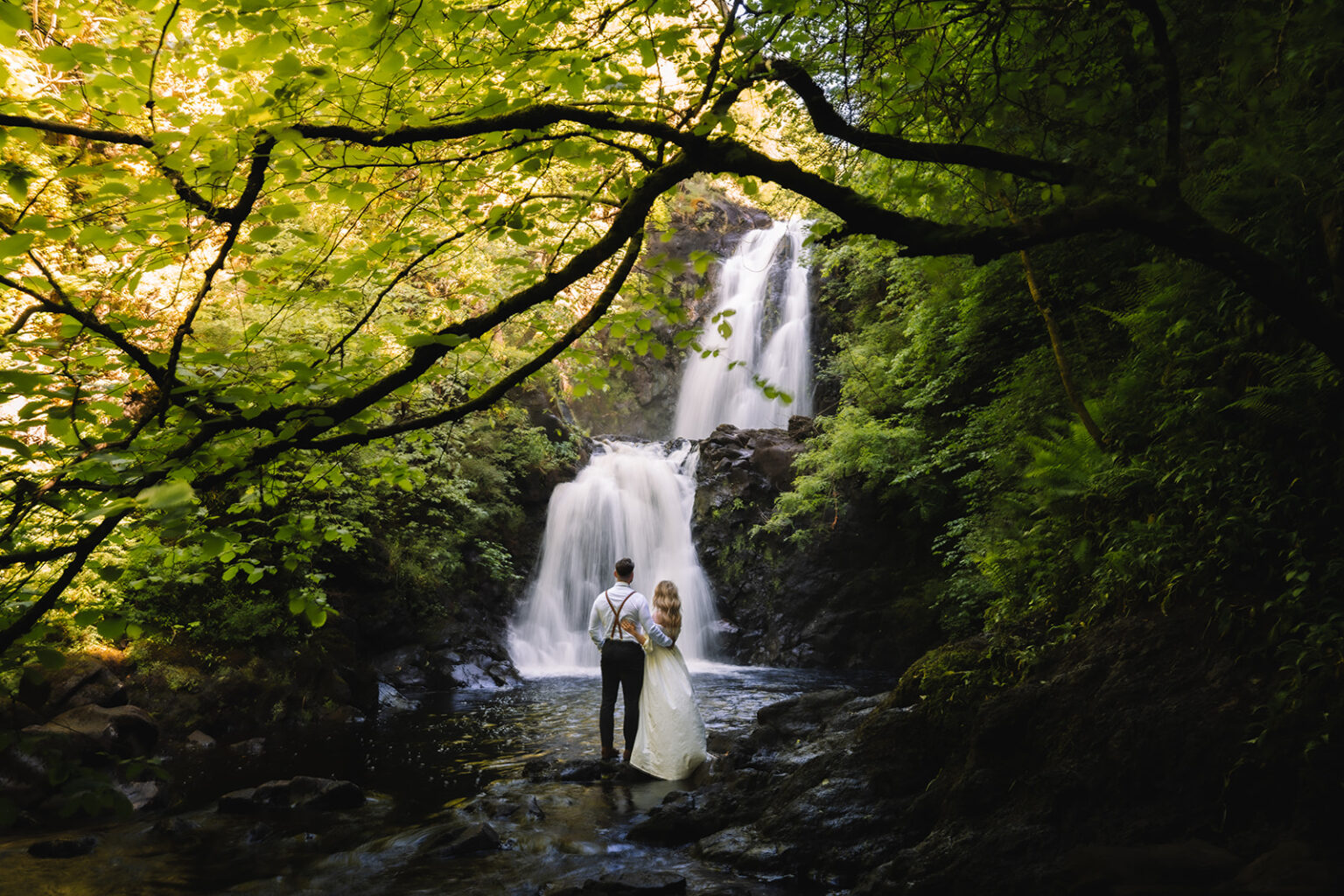 Couple embracing in front of tall forest waterfall.