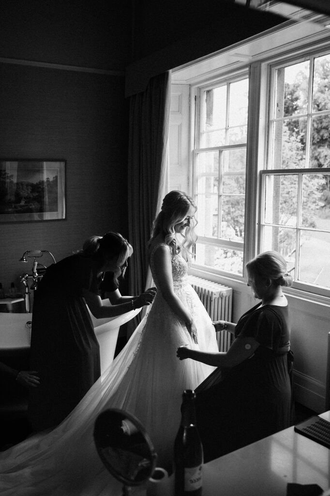 Bride getting ready by window with helper in soft light.