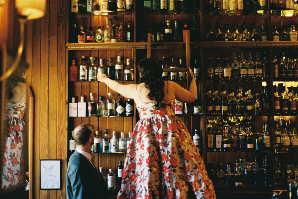 Bride standing on bar counter reaching upward among shelves of bottles.