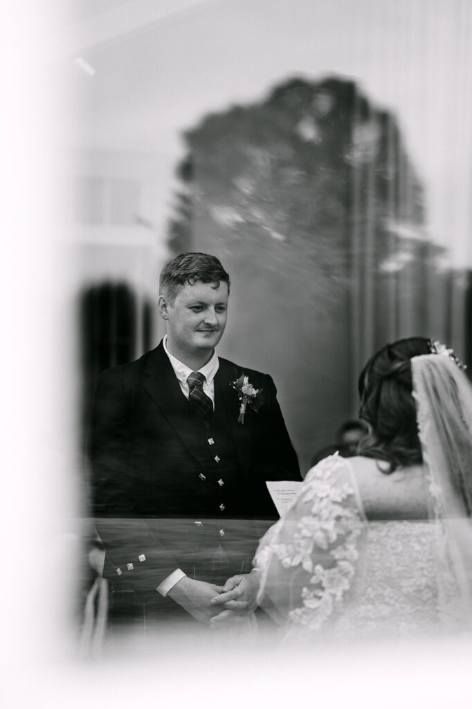 Groom smiling at bride during ceremony seen through soft blur.