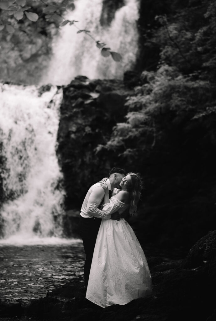 Couple embracing beside waterfall in romantic black and white elopement portrait.