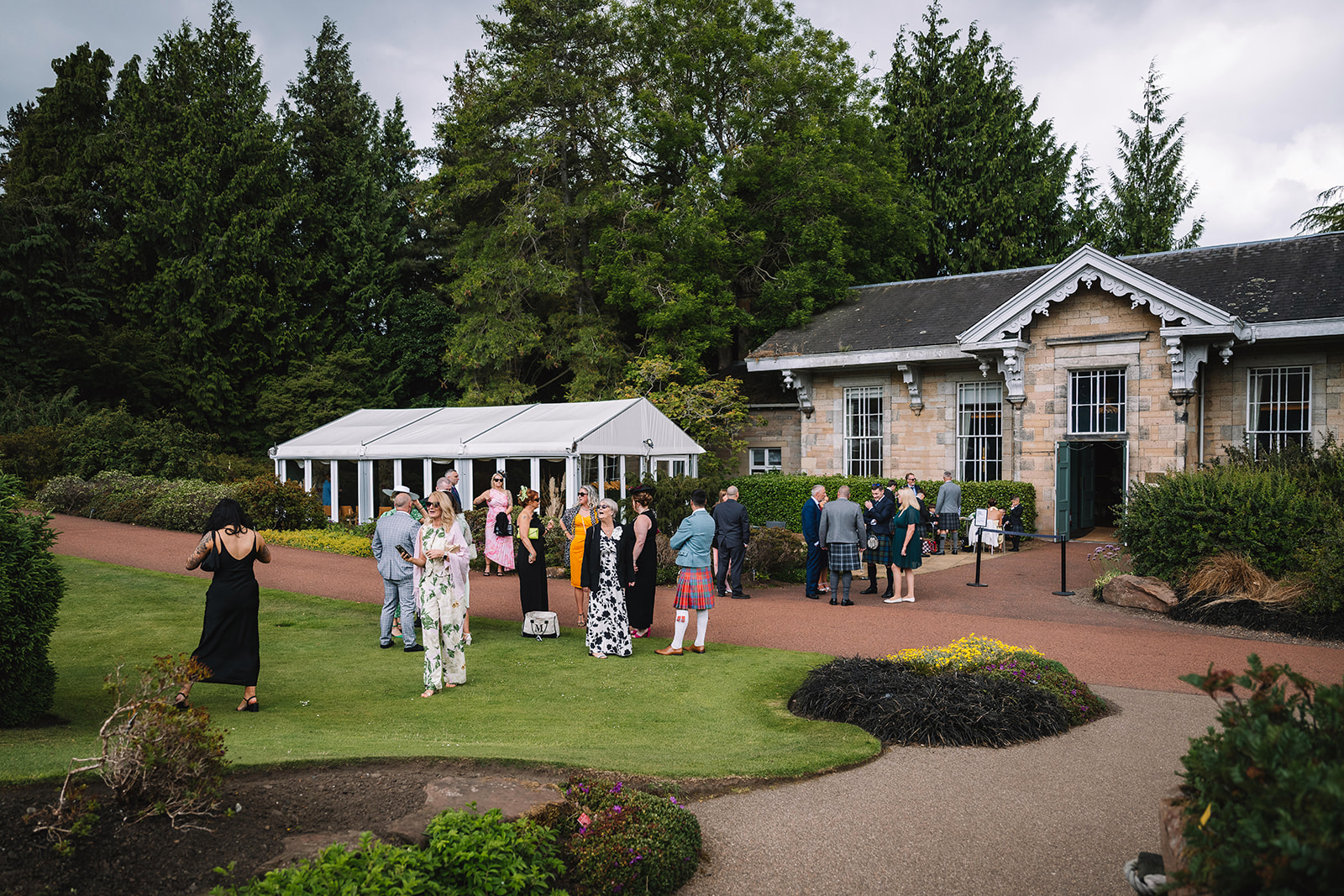 Wedding guests mingling outside venue with garden and buildings.