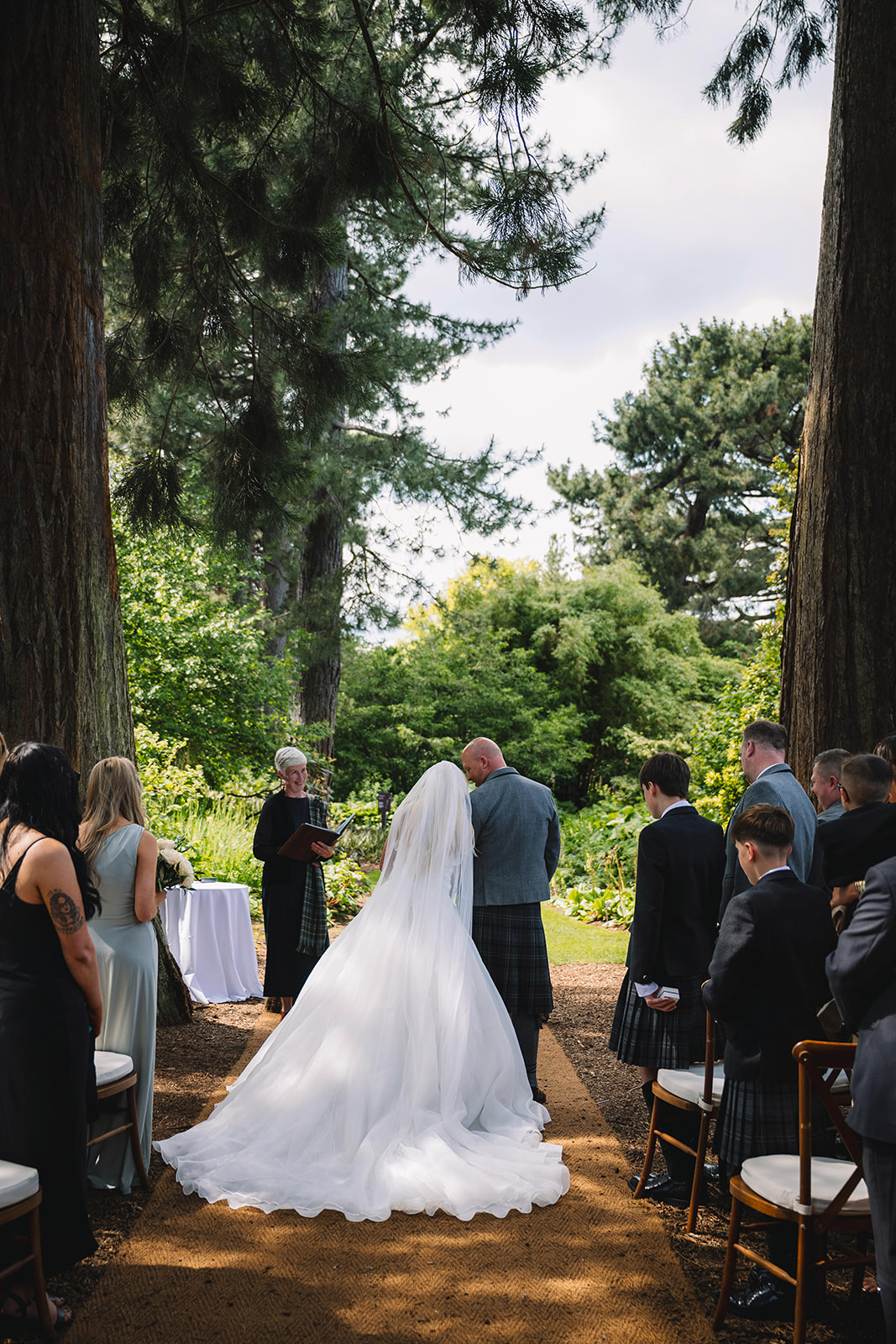 Bride walking down aisle toward ceremony in wooded setting.