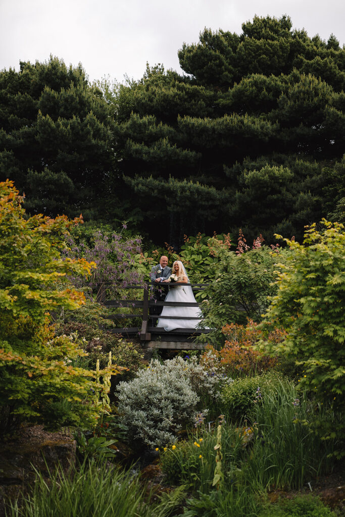 Couple standing on wooden bridge surrounded by lush garden greenery.