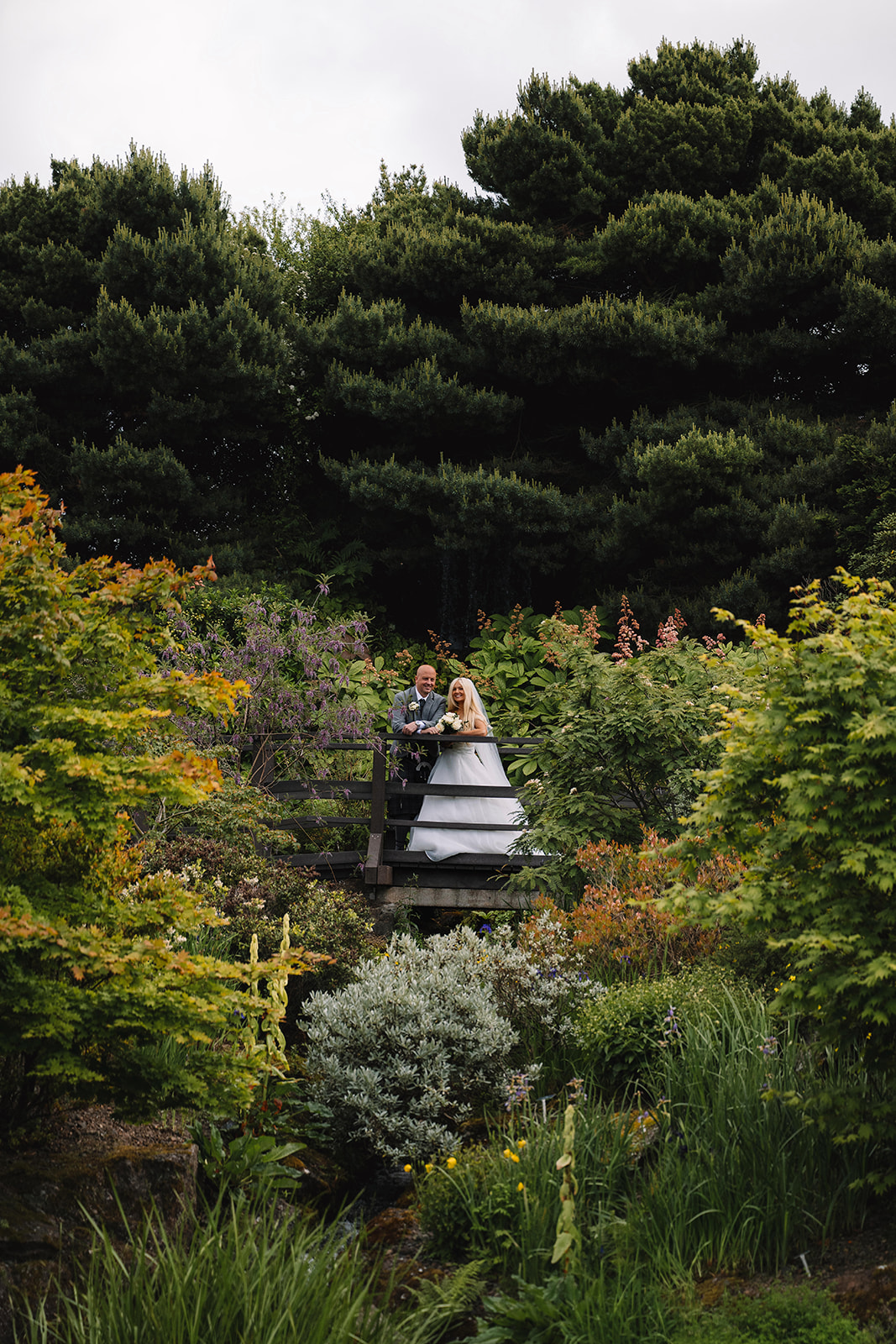 Couple standing on wooden bridge surrounded by lush garden greenery.