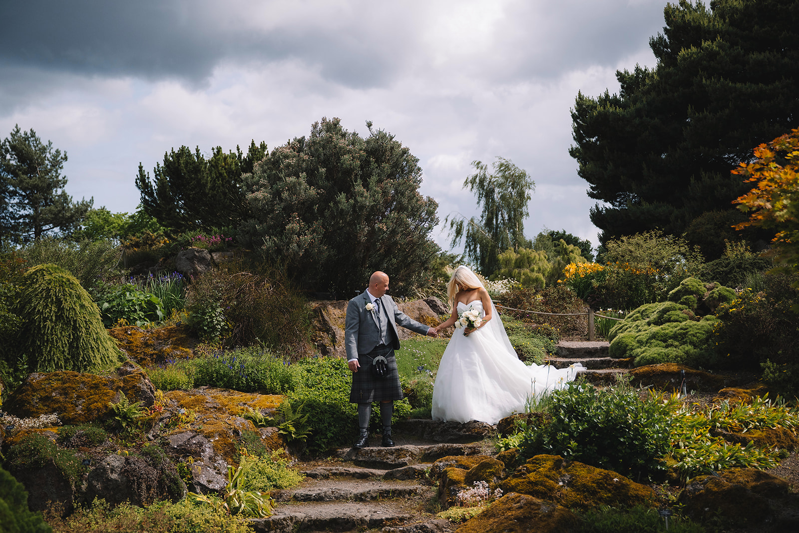 Bride and groom walking down stone steps in landscaped garden setting.