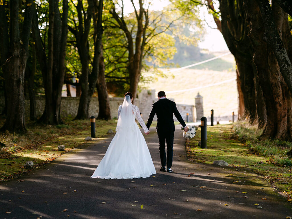 Bride and groom walking hand in hand down tree-lined path.