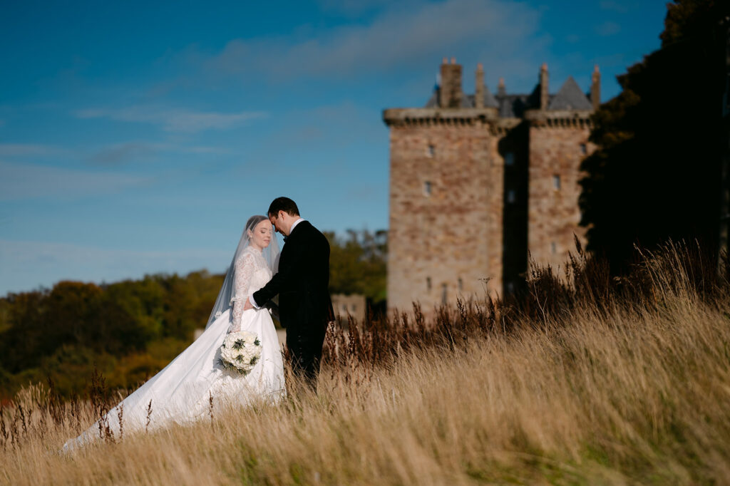Bride and groom embracing near castle ruins in golden grass.