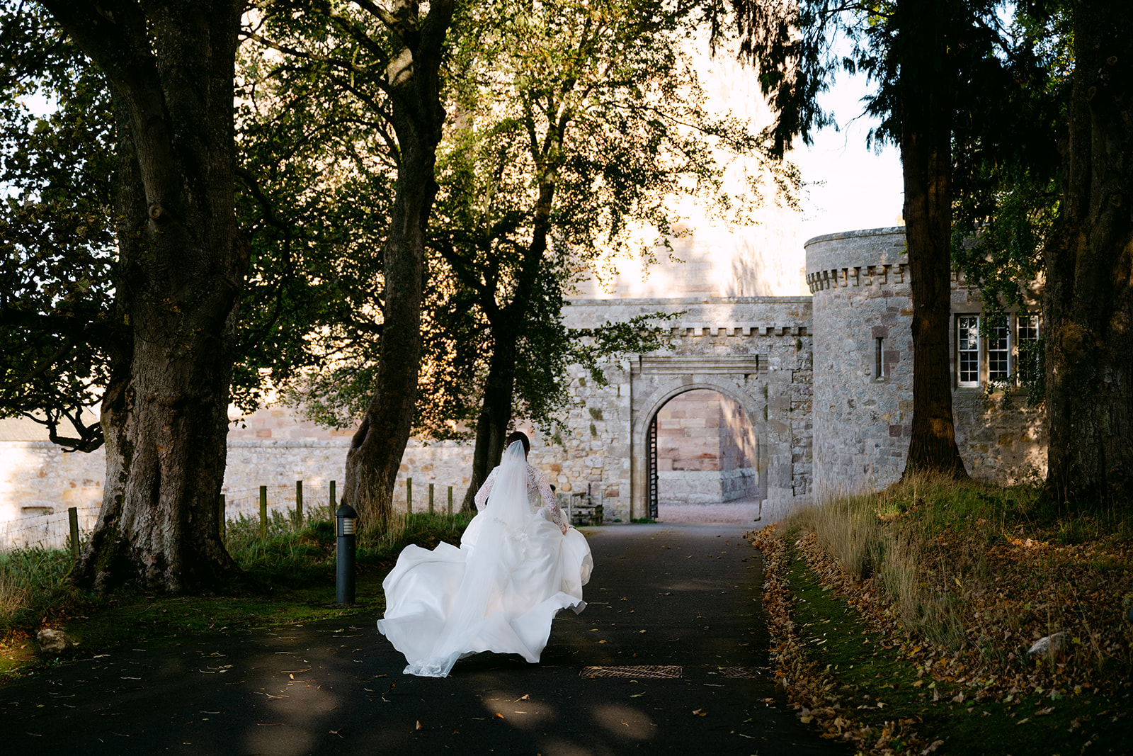 Bride walking toward castle gate through tree-lined path.