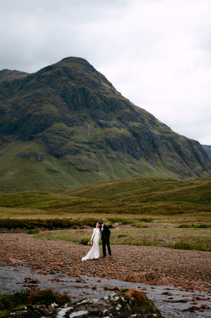 Groom kissing bride’s forehead by a river with Scottish Highlands mountains in the background during intimate wedding.