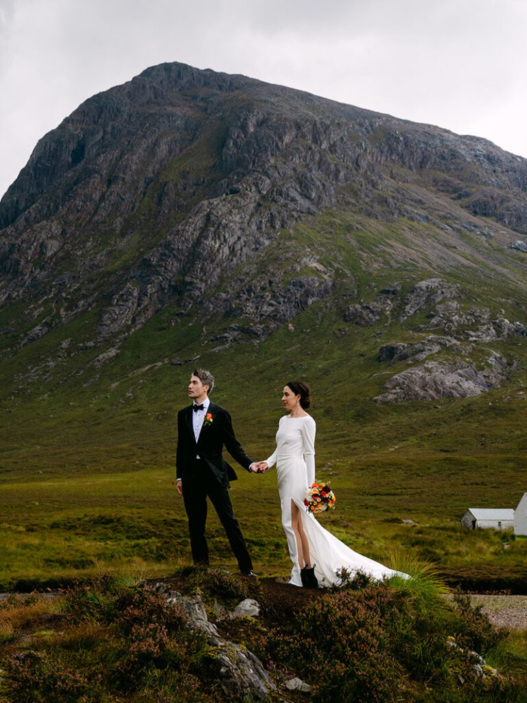 Bride walking toward groom during elopement on grassy hill with Glencoe in background