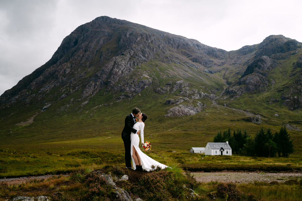 Bride kissing groom during elopement on grassy hill with panoramic glencoe view in background
