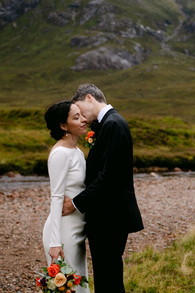 Groom kissing bride’s forehead by a river with Scottish Highlands mountains in the background during intimate wedding.