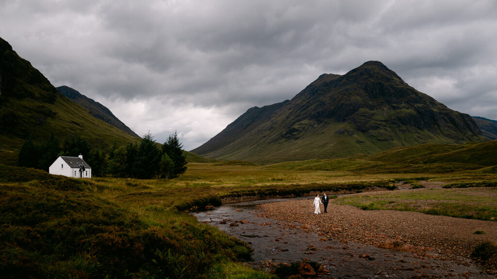 Bride walking toward groom during elopement on grassy hill with panoramic glencoe view in background