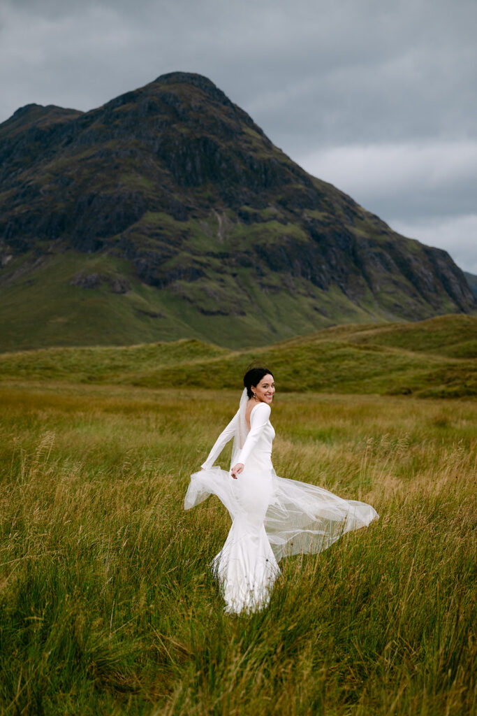 Bride twirling in flowing wedding dress in grassy field with dramatic Scottish Highlands mountain backdrop.